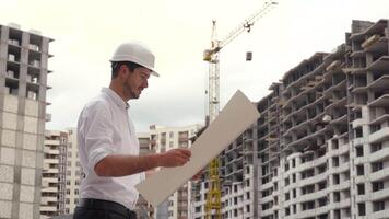 retrato de un exitoso joven ingeniero, arquitecto, constructor, empresario, vistiendo un blanco casco, en un camisa, participación un proyecto en su mano, un rascacielos antecedentes y un construcción sitio video