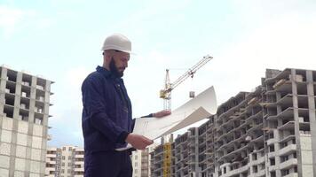 Portrait of a successful young engineer, architect, builder, businessman, wearing a white helmet, in a shirt, holding a project in his hand, a skyscraper background and a construction site video