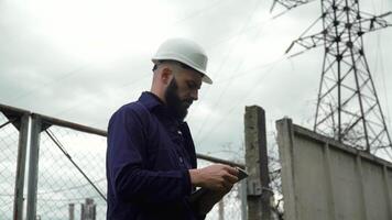 Engineer in special uniform standing with tablet near power station. Male monitoring system performance and making notes in program. Energy efficiency conception video