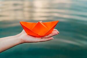 An orange paper ship in a female hand over the water. Woman's hand holding paper boat above the water. Close-up photo