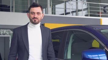 Stylish man is looking into the cam standing near the blue car in car showroom. Technology and people concept video