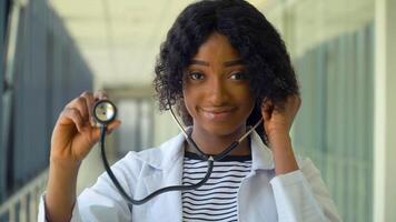 simpático contento africano americano mujer médico o enfermero profesional general facultativo posando con estetoscopio mirando a cámara en médico oficina. sonriente joven hembra médico cerca arriba retrato video