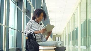 afrikansk amerikan kvinna studerande Sammanträde på de golv med en läsplatta i de universitet. ny modern fullt funktionell utbildning anläggningen. begrepp av uppkopplad utbildning video