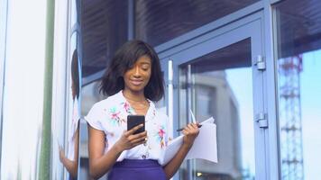 elegante bellissimo africano americano donna d'affari utilizzando sociale reti su sua smartphone e detiene documenti. avendo simpatico aspetto e affascinante Sorridi video