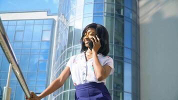 Adult african american young businesswoman is talking on phone outside near modern office building video