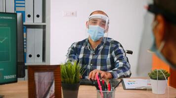 Handicapped businessman with visor and protection mask discussing with colleague working in new normal business office. Man respecting social distance in financial company during global pandemic. photo