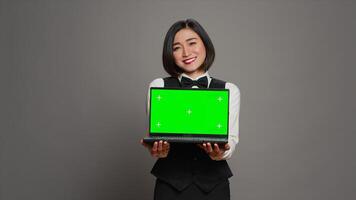 Receptionist showing greenscreen display on personal laptop, presenting isolated copyspace mockup on screen. Asian hotel employee holding pc with chromakey mockup template. Camera A. photo