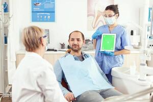 Stomatology nurse pointing her hand on mock up green screen chroma key tablet with isolated display. Sick man patient sitting on dental chair in stomatology office waiting for toothache treatment photo