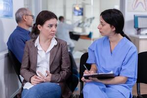 Nurse taking notes on clipboard about patient dental problems waiting for orthodontist sitting on chair in waiting room of stomatological clinic. Assistant explaining medical procedure to woman. photo