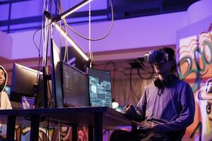 Hacker wearing vr glasses, immersed in simulation while doing illegal activity. Young criminal using virtual reality headset to code malicious software in metaverse in abandoned warehouse photo