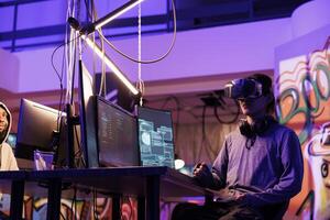 Hacker wearing vr glasses, immersed in simulation while doing illegal activity. Young criminal using virtual reality headset to code malicious software in metaverse in abandoned warehouse photo