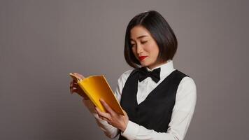 Asian receptionist reading a literature book over grey background, enjoying lecture hobby with a novel story in studio. Hotel concierge employee reads a fiction tale for knowledge. Camera B. photo