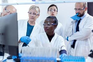 Multiethnic group of laboratory scientists discussing their research in front of computer. African healthcare researcher in biochemistry laboratory wearing sterile equipment. photo