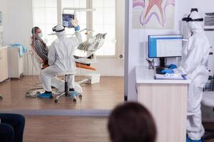 Stomatologist in protective suit asking for patient dental x-ray examining teeth problems during coronavirus pandemic in modern clinic. Medical team wearing coverall, face shield, mask and gloves. photo