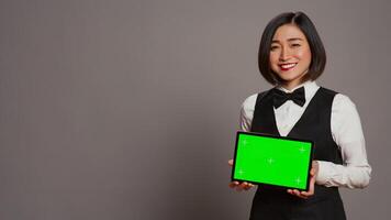 Asian receptionist holding tablet with greenscreen on camera, presenting mockup template on modern gadget. Hotel concierge looking at copyspace layout display, grey studio. Camera B. photo