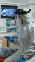 Senior woman showing aching tooth to dentist dressed in coverall during coronavirus epidemic. Assistant and dentist doctor talking with patient wearing protective suit, face shield, mask and gloves photo