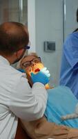 ortodoncista Encendiendo el lámpara Hasta que examen y paciente apertura boca. estomatólogo Hablando a mujer con dolor de muelas sentado en estomatológico silla mientras enfermero preparando herramientas para cirugía. foto