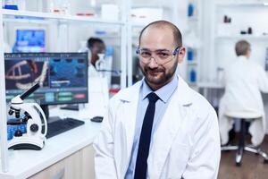 farmacéutico investigador sentado en taburete mirando a camara.serio experto en genética en laboratorio con moderno tecnología para médico investigaciones con africano asistente en el antecedentes. foto