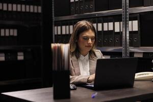 Bookkeeper analyzing administrative files on laptop computer, searching for bureaucracy record in corporate depository. Manager in formal suit working at accountancy report in storage room photo