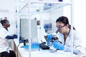 Expert in in genetics doing research using microscope and african assistant in the background. Chemist wearing lab coat using modern technology during scientific experiment in sterile environment. photo