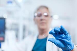 Close up of senior scientist holding biochemistry material on glass slide during clinic study. Elderly researcher in sterile lab looking on microscope slide wearing lab coat. photo