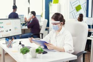 Entrepreneur wearing face mask against covid19 using wireless headphones for remote call. Employees with visors working in corporate company workspace respecting social distance analysing data and graphs. photo