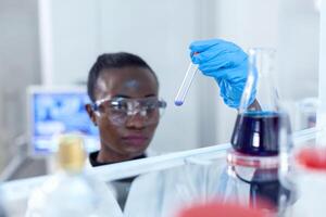 African scientist looking pensive at test tube with blue liquid. Black researcher in sterile laboratory conducting pharmacology experiment wearing coat. photo