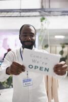 Fashion shop happy african american man assistant hanging opening sign on boutique window. Smiling cheerful clothing store employee holding sign board noticing buyers about work day beginning photo