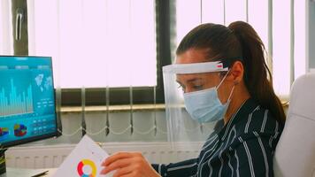 Close up of businesswoman wearing protection mask and visor checking annual financial reports working in new normal business office respecting social distance. Freelancer using modern technology photo