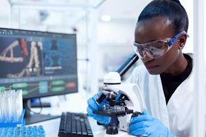 African pharmacology researcher analysing chemical substance using microscope for study.. Black healthcare scientist in biochemistry laboratory wearing sterile equipment. photo