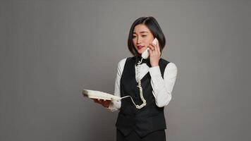 Asian receptionist answering landline phone call in studio, talking on vintage telephone with cable. Woman front desk staff using phone with cord to confirm room reservation. Camera A. photo