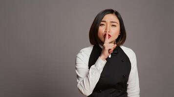 Restaurant hostess showing silence symbol in studio, doing hush mute gesture with finger over lips. Private waitress asking to keep secret with confidential information, secrecy sign. Camera B. photo