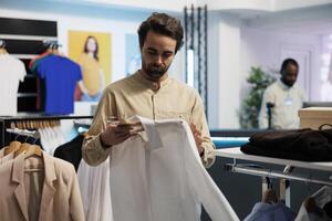 hombre comprobación elegante casual camisa en compras centro comercial Departamento salida. joven cliente examinando de moda prenda en percha mientras elegir vestir en Al por menor centrar Moda boutique foto