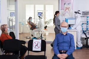 Dentist nurse dressed in ppe suit with face shiled discussing with patient in stomatology waiting room. People keeping social distancing as prevention during coronavirus outbreak. photo