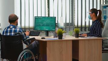 Woman entrepreneur bringing immobilized businessman at workplace with wheelchair wearing protection visor working in new normal business office. Businesspeople with masks respecting social distance photo