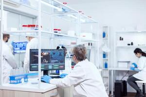 Elderly aged medical chemist working on powerful computer in modern facility. Senior scientist in pharmaceuticals laboratory doing genetic research wearing lab coat with team in the background. photo