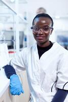 African scientist wearing protective glasses in medical laboratory looking at camera. Multiethnic team of researchers working in microbiology lab testing solution for medical purpose. photo