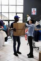 Tienda trabajador explicando cliente Envío detalles a africano americano mensajero en moderno boutique. hombre vistiendo entrega uniforme, participación cartulina cajas con en línea pedidos en compras centrar foto