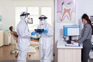 Stomatology team dressed up in ppe suit during global pandemic with coronavirus in dental reception holding patient x-ray, keeping social distancing. Receptionist with face mask as prevention. photo