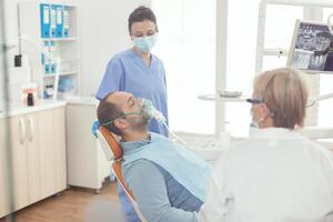 hospital asistente poniendo oxigeno máscara antes de dental cirugía sentado en estomatología silla en odontología clínica . mayor ortodoncista médico y enfermero trabajando en moderno oficina vistiendo máscara y guantes foto