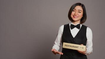 Asian hotel concierge holding restaurant sign to indicate direction, pointing towards dining area. Receptionist assisting clients to enjoy all amenities, stands over grey background. Camera B. photo