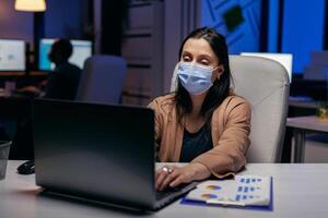 sobrecargado de trabajo mujer de negocios vistiendo cara máscara como la seguridad prevenido debido a empresa reglas. mujer siguiendo social distanciamiento reglas debido a coroanvirus pandemia mientras trabajando tarde horas a el oficina. foto