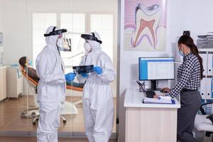 Dentist assistant discussing with doctor patient diagnosis keeping social distancing dressed in ppe suit face shiled, during global pandemic with coronavirus holding x-ray. photo