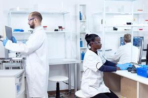 African scientist working on computer wearing protection gear doing analysis. Multiethnic team of researchers working in microbiology lab testing solution for medical purpose. photo