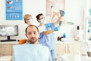 frente ver de enfermo hombre mirando dentro cámara mientras esperando médico equipo a examinando somatología radiografía sentado en dental silla en hospital deudor oficina. estomatólogo haciendo diente cita foto