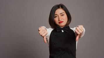 Asian restaurant hostess showing thumbs down and dislike sign, expressing negativity and disapproval in studio. Displeased waitress working at a diner and serving customers. Camera B. photo