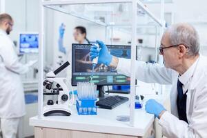 Experienced scientist working in busy laboratory at his workplace holding dropper dispense. Senior professional chemist using pippete with blue solution for microbiology tests. photo