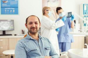 Man patient looking into camera waiting for radiography results sitting on dental chair in hospital stomatology clinic. In background dentists team looking at tooth x-ray preparing for surgery photo