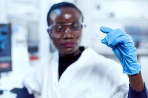 Multiethnic researcher during study holding microscope sample on glass slide. African healthcare scientist in biochemistry laboratory wearing sterile equipment. photo