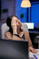 cansado mujer de negocios acuerdo ojos cerrado frotamiento frente porque agotador. empleado que cae dormido mientras trabajando tarde a noche solo en el oficina para importante empresa proyecto. foto